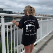 Person wearing a black t-shirt with 'ANTI-SOCIAL DOG OWNER' text and graphic, standing on a bridge.