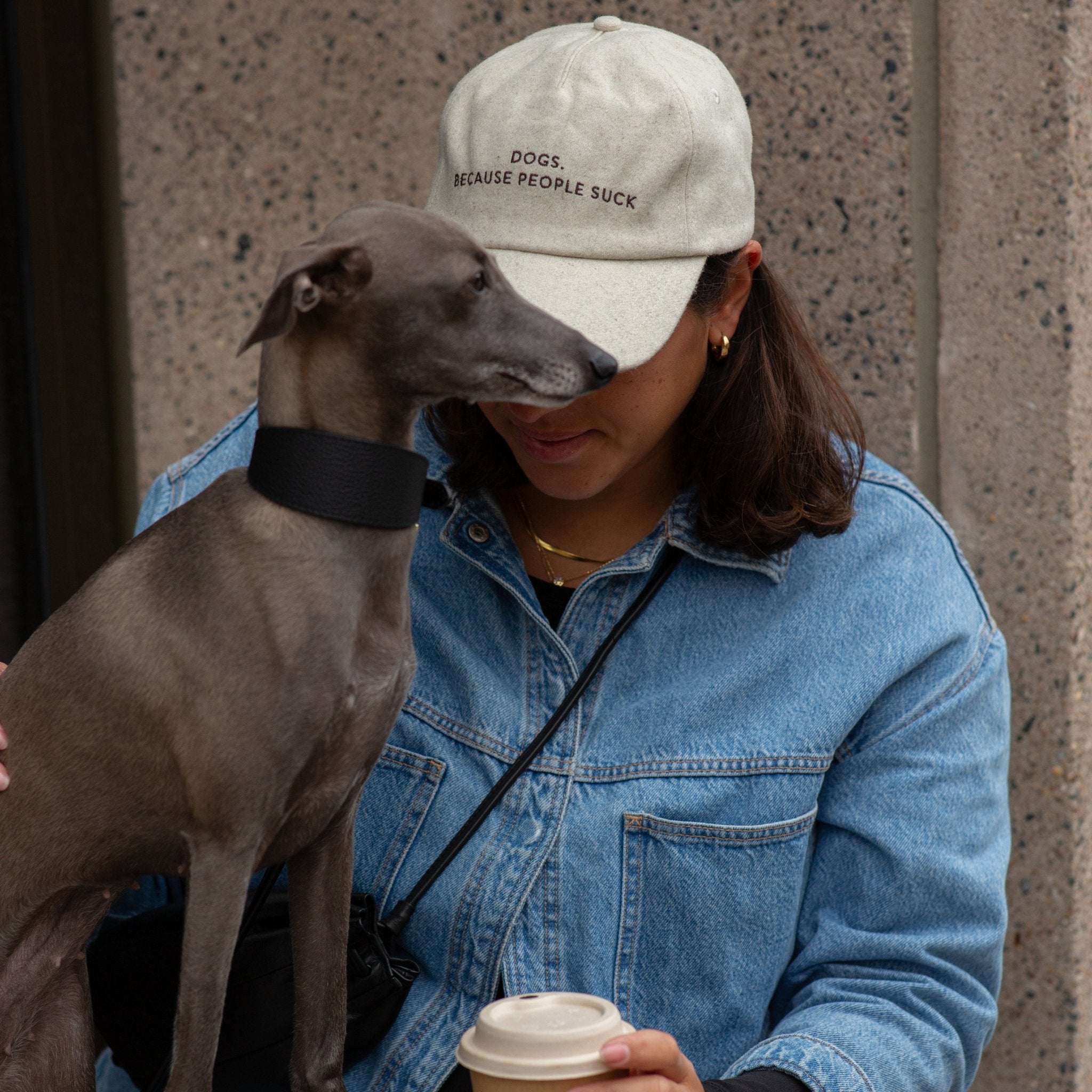Person wearing a cap with text, holding a dog and a coffee cup, against a concrete wall.